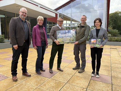 Fünf Personen stehen vor einem Gebäude und lächeln in die Kamera. Zwei von ihnen halten einen großen Spendenscheck und ein Buch.